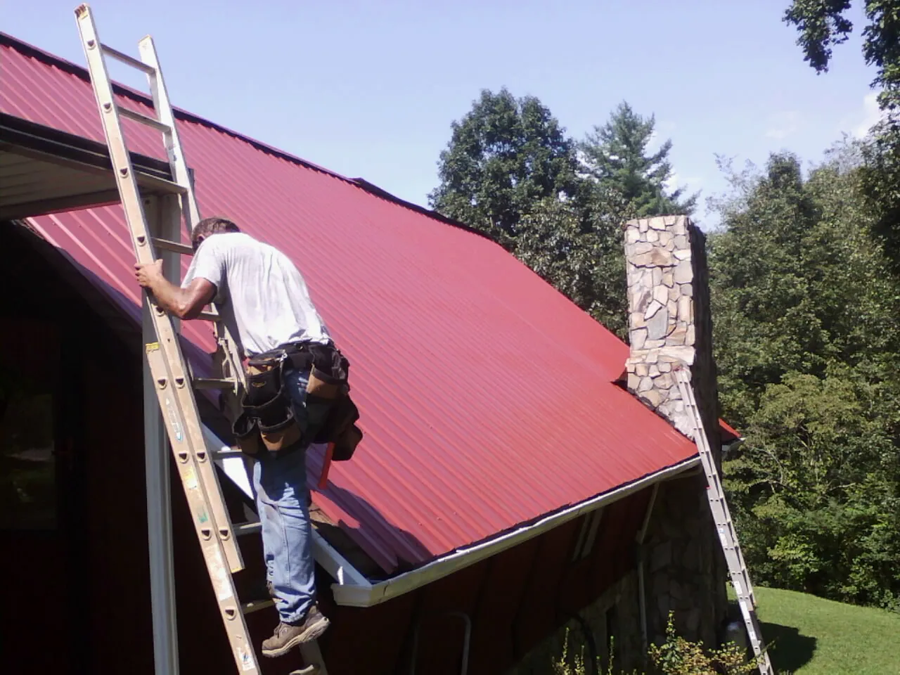 Roofer on a ladder beside a red metal roof and stone chimney — Soffit & Fascia Repair by Roofing Brothers in New Rochelle